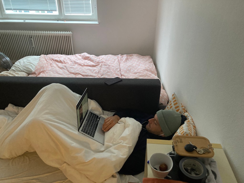 A person lying on a couch with a laptop, wrapped in a blanket, in a cozy indoor setting. A bed with pink bedding is visible in the background, along with a small table containing a cup, keys, and other items.